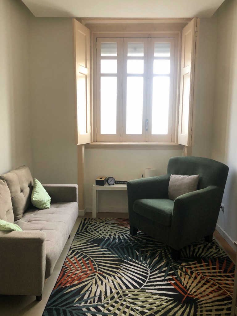 Sala de consultas de um dos espaços da associação casa estrela do mar em Berna. Sofa de casal cinzento/creme, em frente um sofa singular verde escuro. Por baixo um tapete com motivos naturais (folhas de árvores).